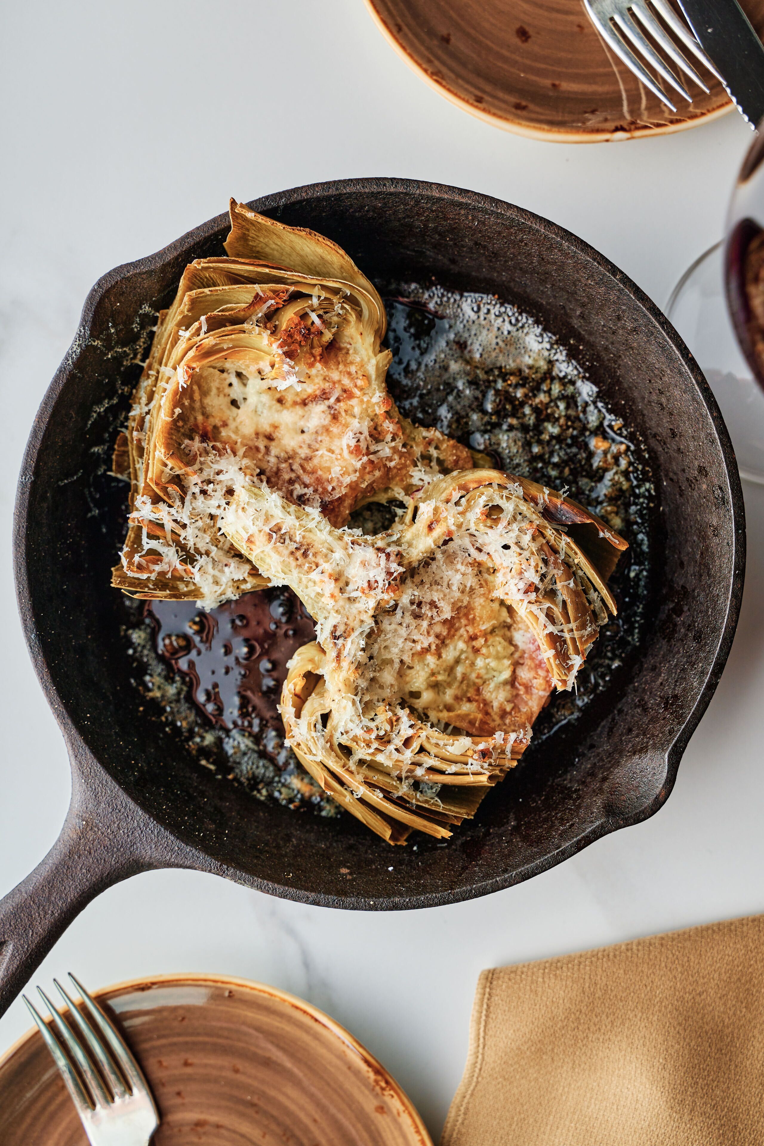 Une poêle en fonte contenant un plat, vraisemblablement du poulet cuit au four, garni de fromage et d’herbes, posé sur une surface blanche.