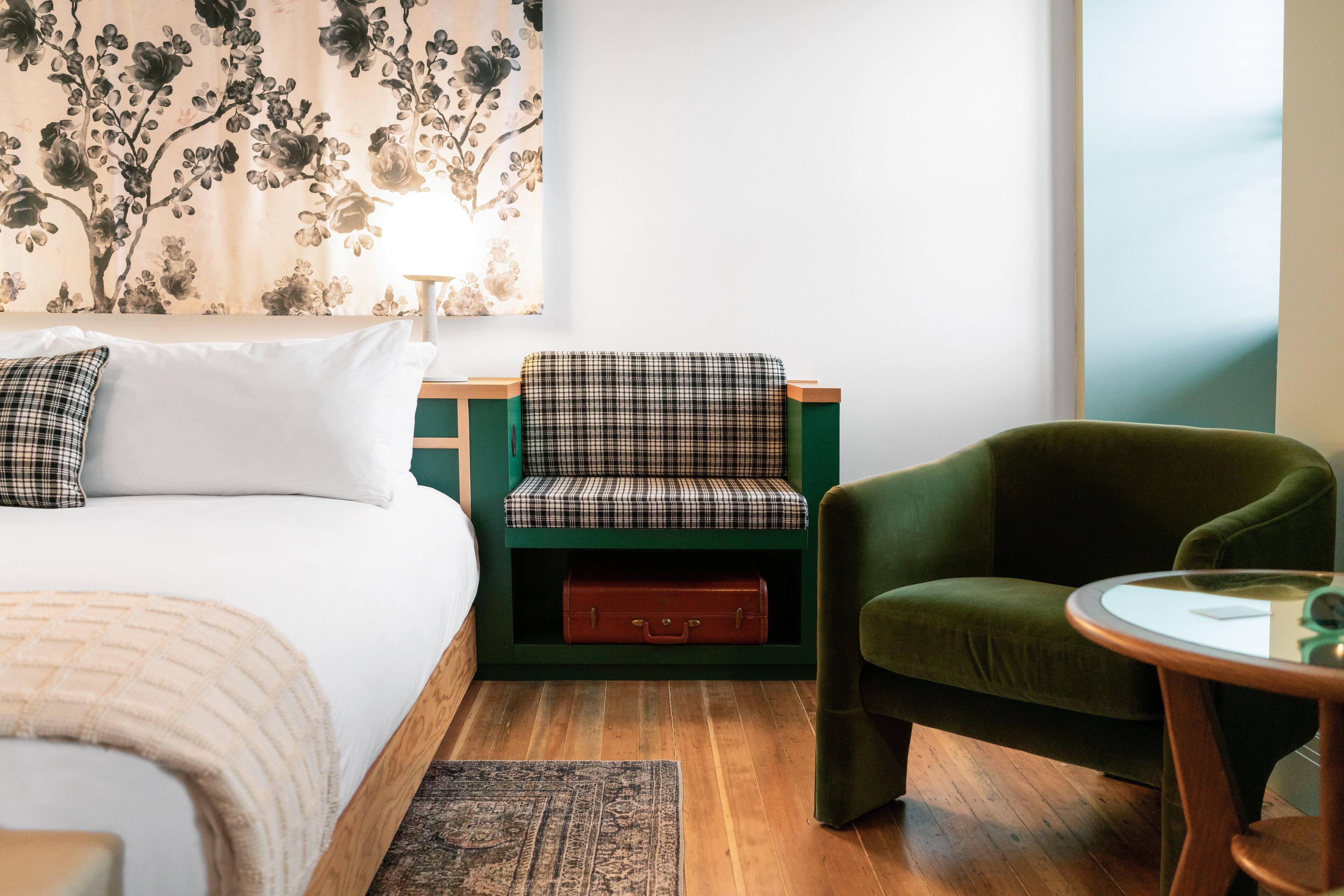 A hotel room with a bed, a green armchair, a small table, a bench with a suitcase, and a floral wall tapestry.
