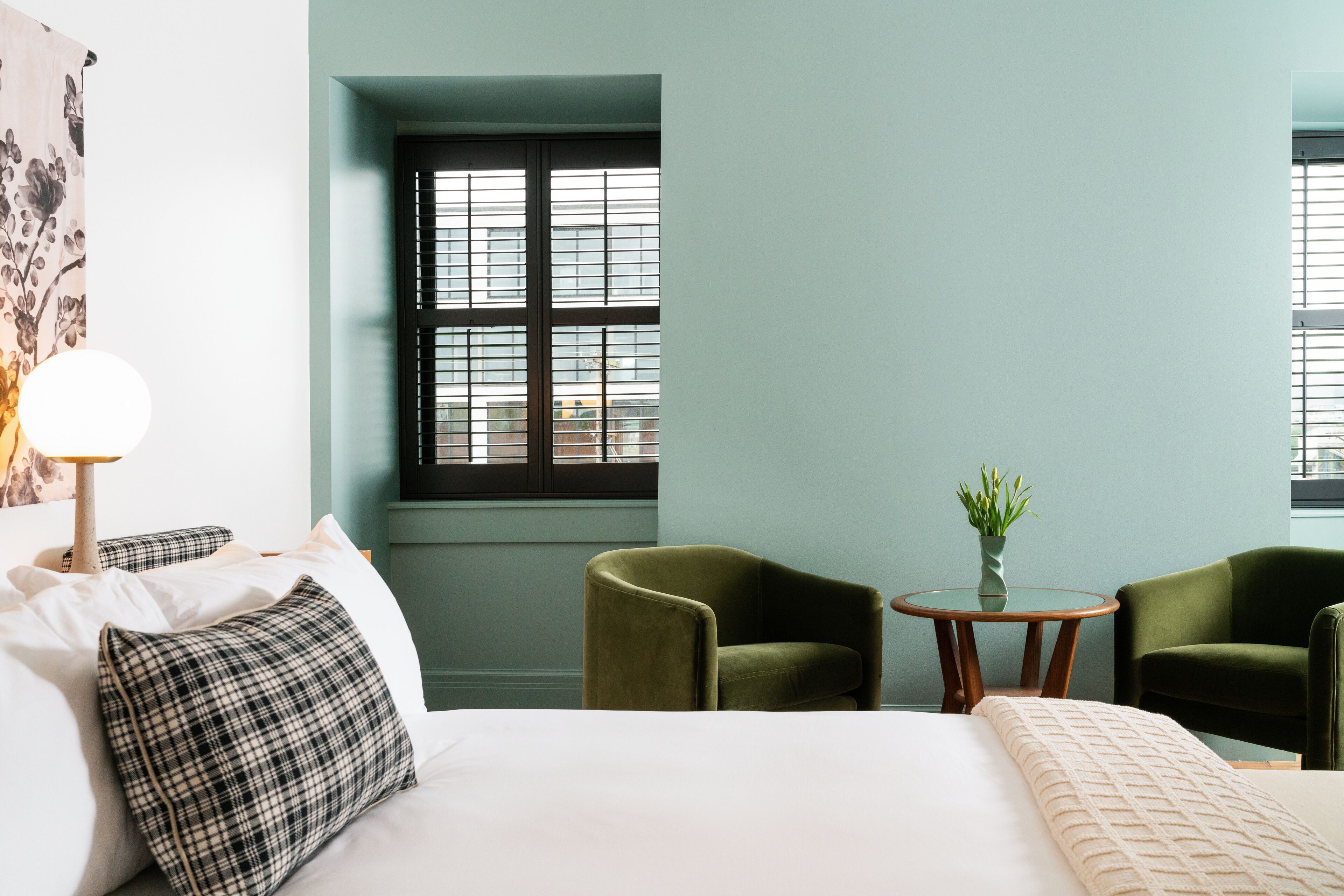 A hotel room with a bed, two green armchairs, a small round table with a vase of flowers, and a window with blinds.
