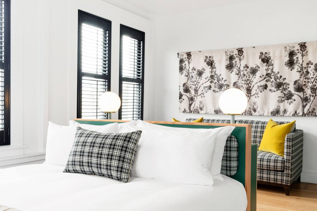 A modern bedroom with a large bed, a desk, and a chair. There are two windows with blinds, a wall with a floral tapestry, and a hanging light fixture.