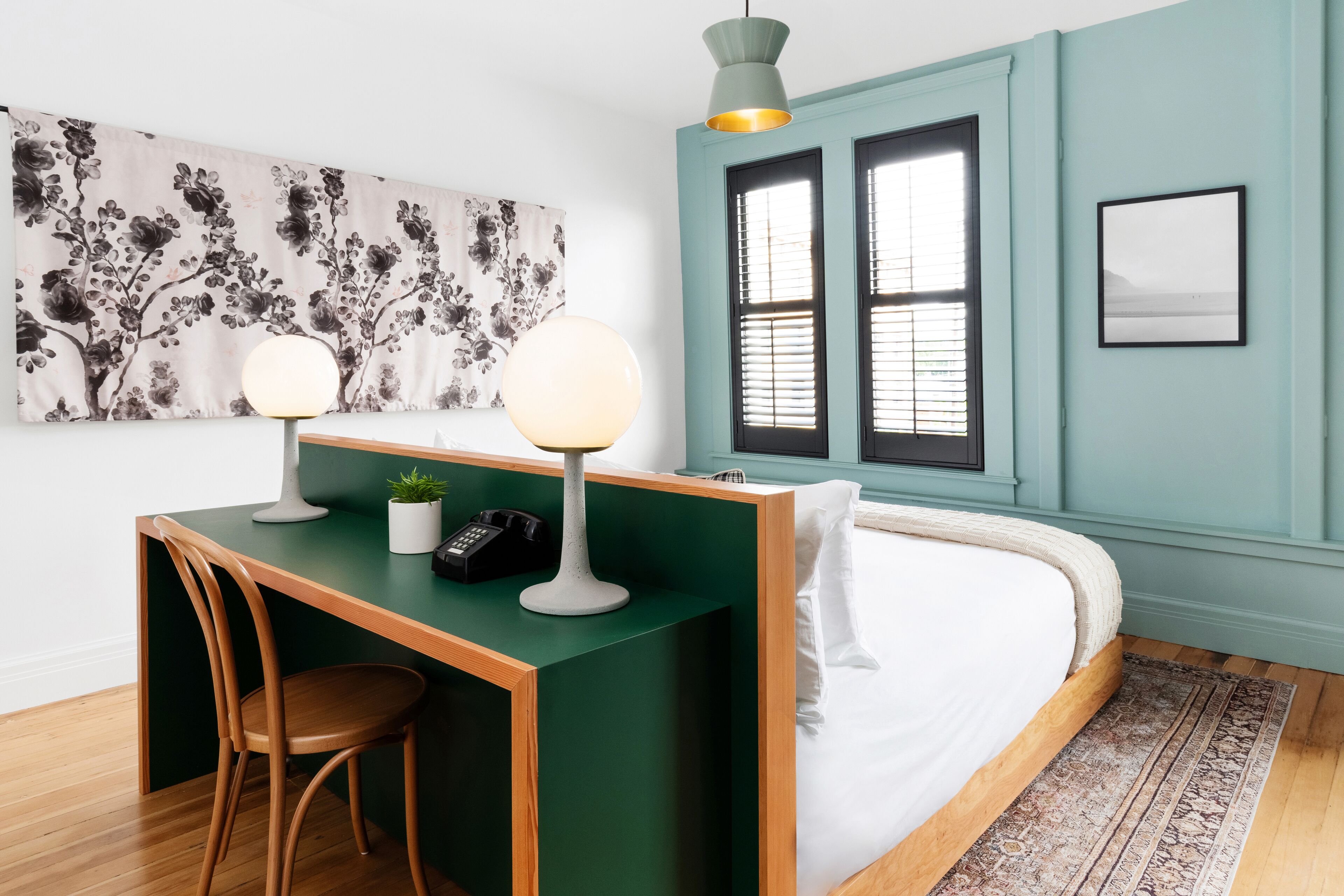 A room with a bed, a desk with two lamps, a chair, a plant, and a painting on the wall.