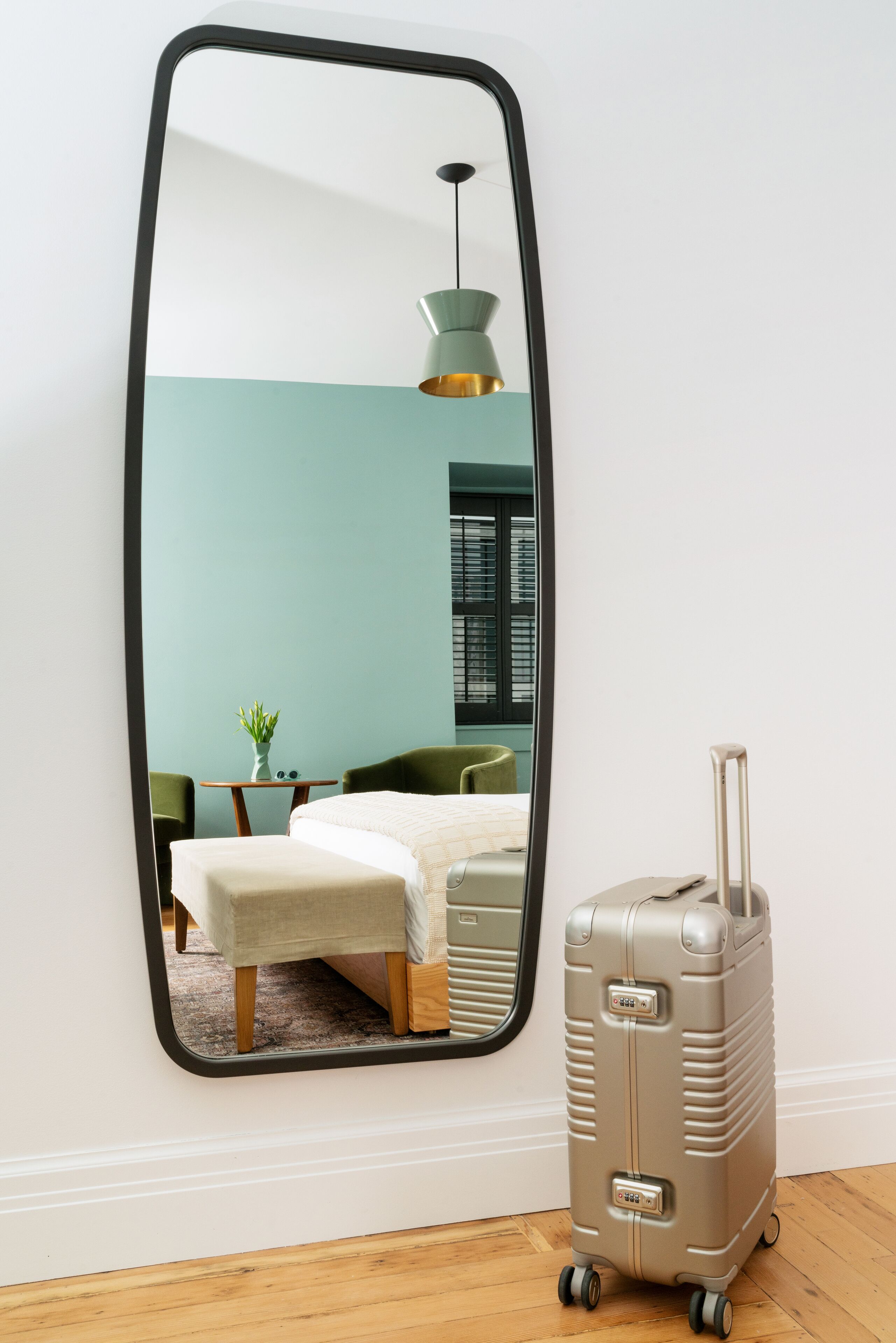 A room with a large mirror, a bed, a green chair, a small table with a plant, and a suitcase.