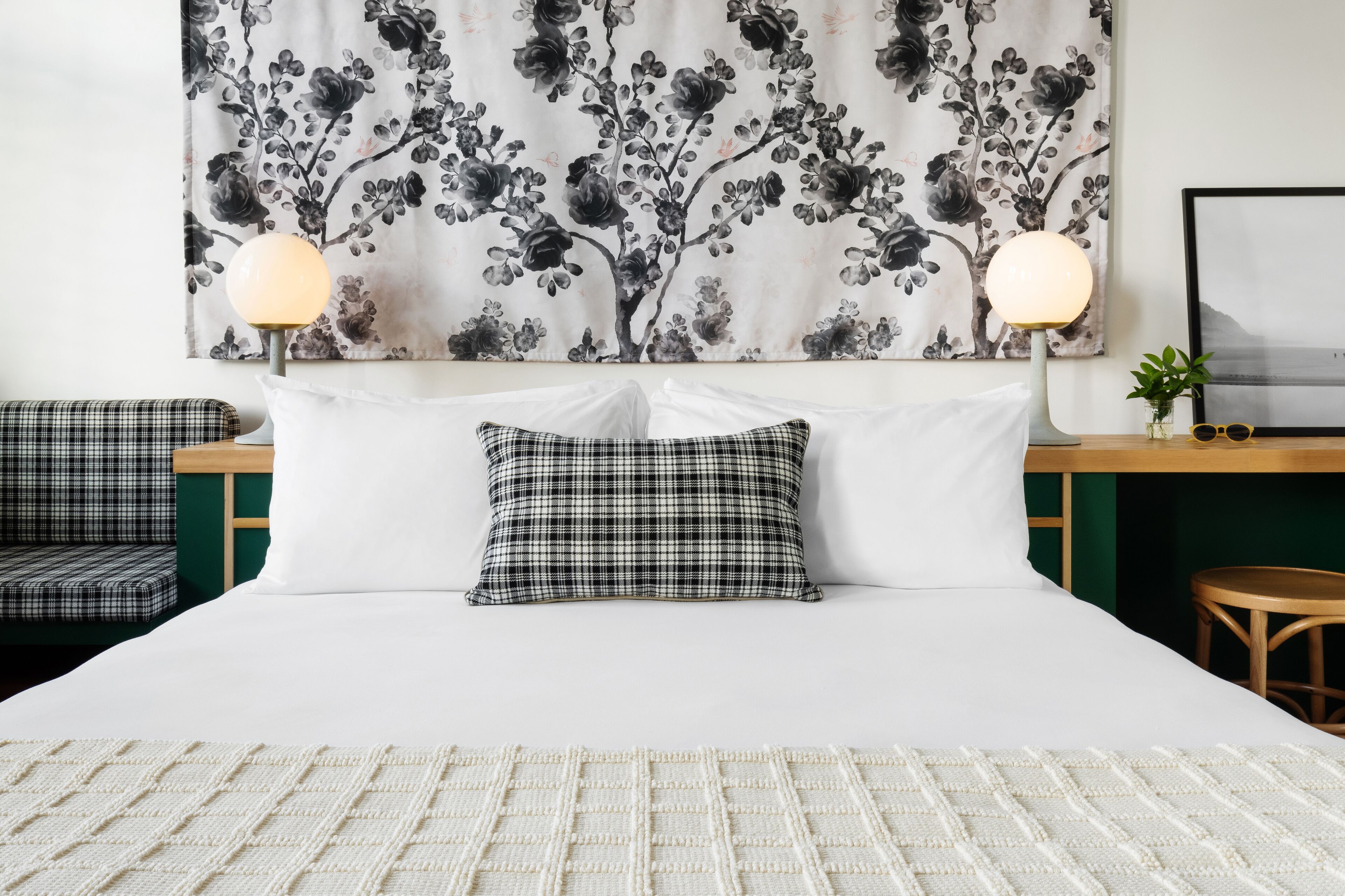 Ein ordentlich eingerichtetes Hotelzimmer mit einem großen Bett, zwei Nachttischlampen, einem karierten Kissen, einem gerahmten Bild an der Wand und einer kleinen Pflanze auf dem Nachttisch.