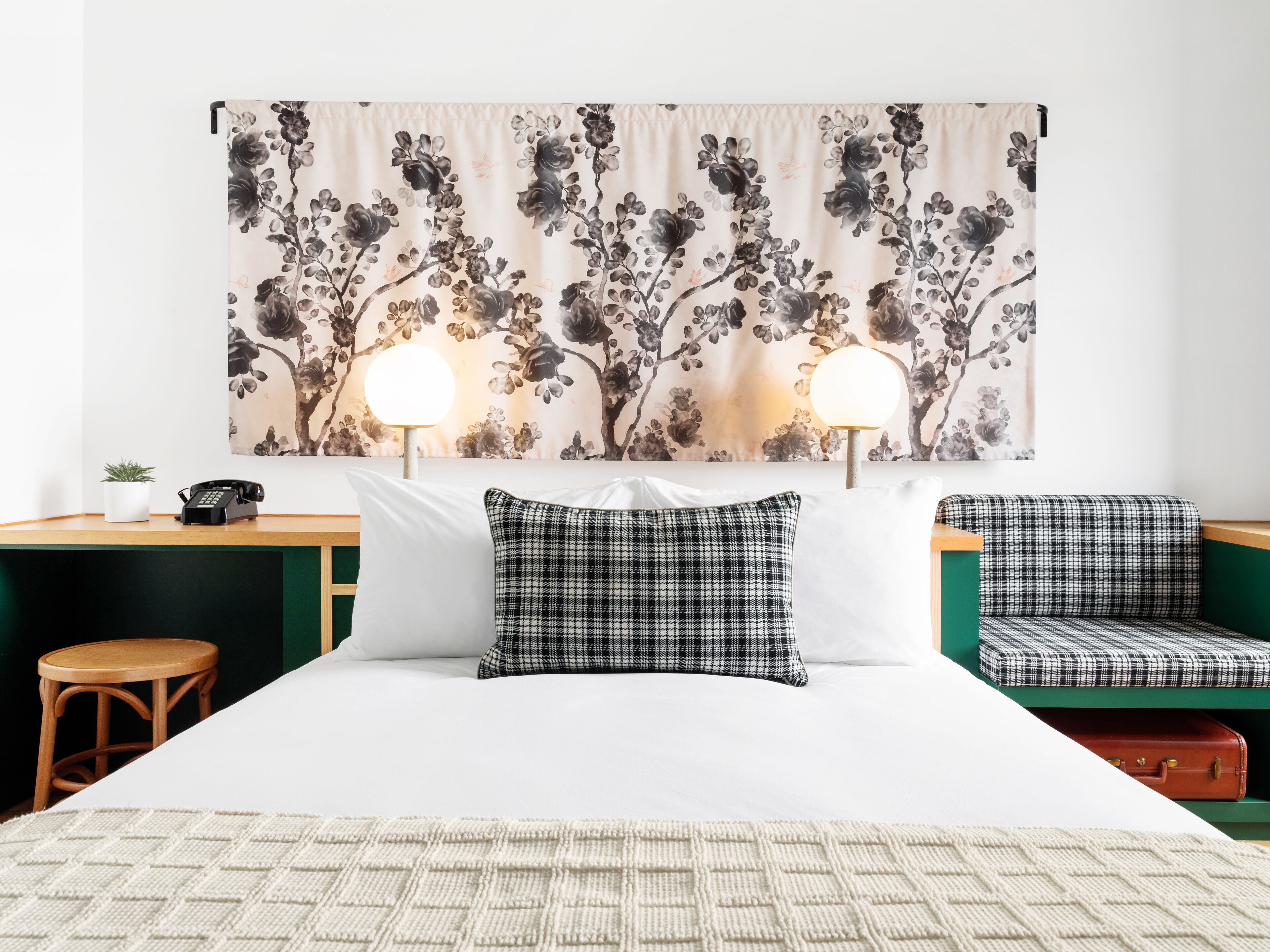 A hotel room with a bed, two bedside tables, a desk, a chair, a lamp, and a suitcase.