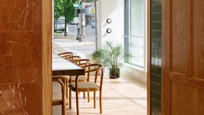 A modern interior with a wooden dining table and chairs, a potted plant, and large windows offering a view of the street outside.