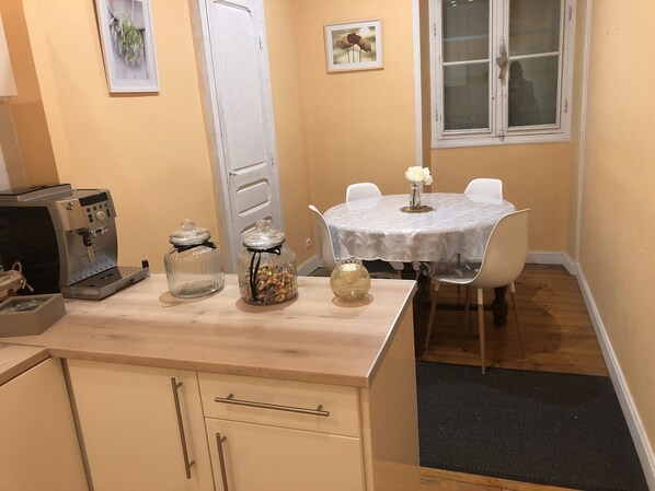 Una pequeña cocina con una cafetera, dos frascos de vidrio, un jarrón con flores y una mesa de comedor con sillas.