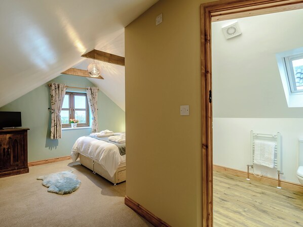 A bedroom with a sloped ceiling, a bed with white linens, a wooden wardrobe, a small rug, and a window with curtains.