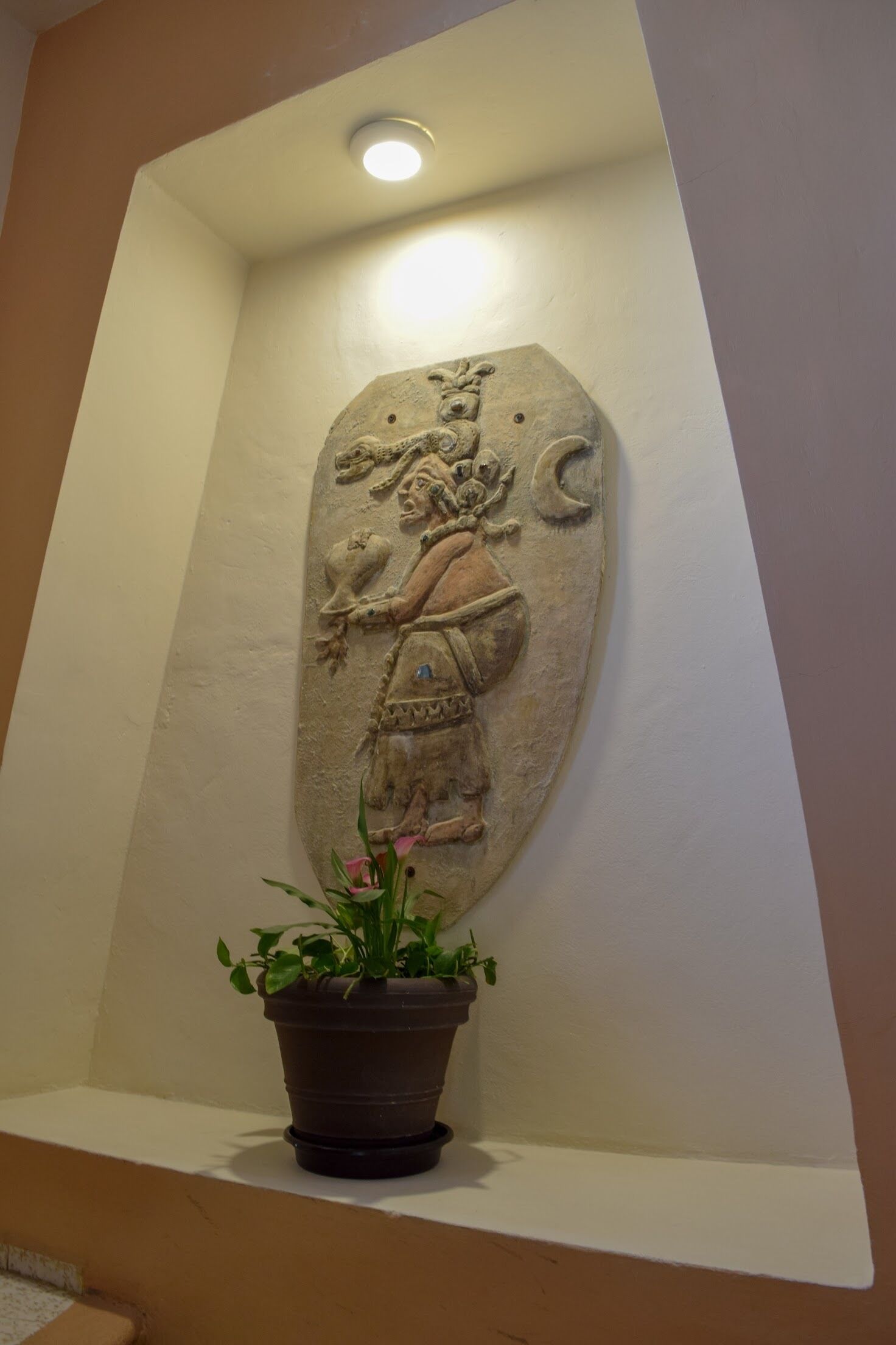 Een reliëfbeeldhouwwerk van steen in een muur, een plant in pot en een plafondlamp.