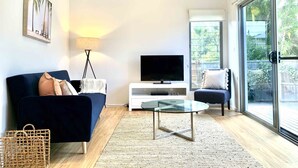 A modern living room with a dark blue sofa, a glass coffee table, a flat-screen TV on a white stand, and a large sliding glass door leading to a balcony with greenery. - Marie’s Byron Escape (Byron Bay)