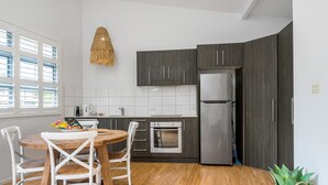 A modern kitchen with dark cabinets, a stainless steel refrigerator, a wooden dining table with chairs, and a hanging light fixture. - Marie’s Byron Escape (Byron Bay)