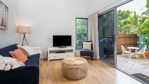 A modern living room with a dark blue sofa, a flat-screen TV on a white console, a wicker ottoman, and a glass door leading to a patio with outdoor furniture. - Marie’s Byron Escape (Byron Bay)