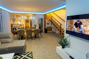 Un salon moderne comprenant un coin repas, un escalier et un téléviseur diffusant un match.
