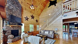 A spacious living room with a stone fireplace, wooden ceiling, and a staircase leading to an upper level. - Bear's Eye View by Jackson Mountain Rentals (Gatlinburg)