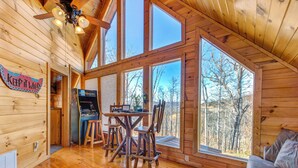 A wooden cabin interior with a dining area, a gaming table, and a large window overlooking a forest. - Bear's Eye View by Jackson Mountain Rentals (Gatlinburg)
