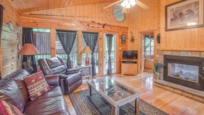 L’intérieur d’un chalet confortable, avec des murs en bois, un canapé en cuir, une table basse en verre, une cheminée et un ventilateur de plafond.