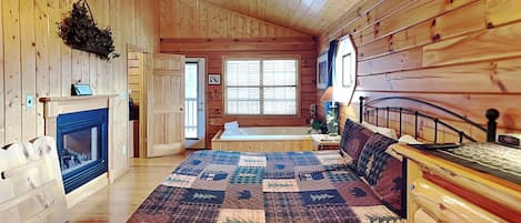 A cozy log cabin bedroom with a large bed, a fireplace, a ceiling fan, and a window with blinds.