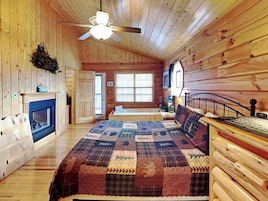 A cozy log cabin bedroom with a large bed, a fireplace, a ceiling fan, and a window with blinds.