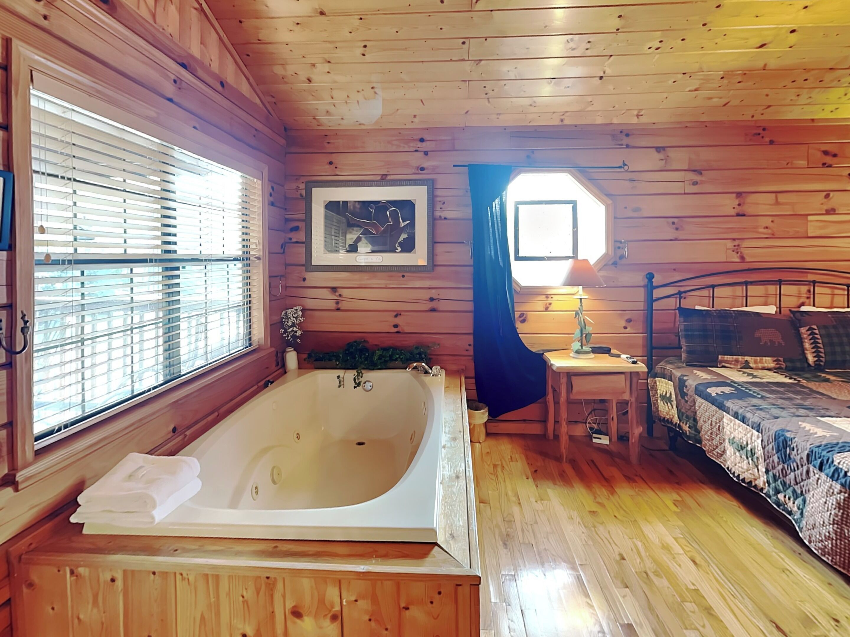 A wooden cabin bedroom with a large bathtub, a bed with a quilt, and a small table with a lamp.