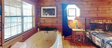 A wooden cabin bedroom with a large bathtub, a bed with a quilt, and a small table with a lamp.