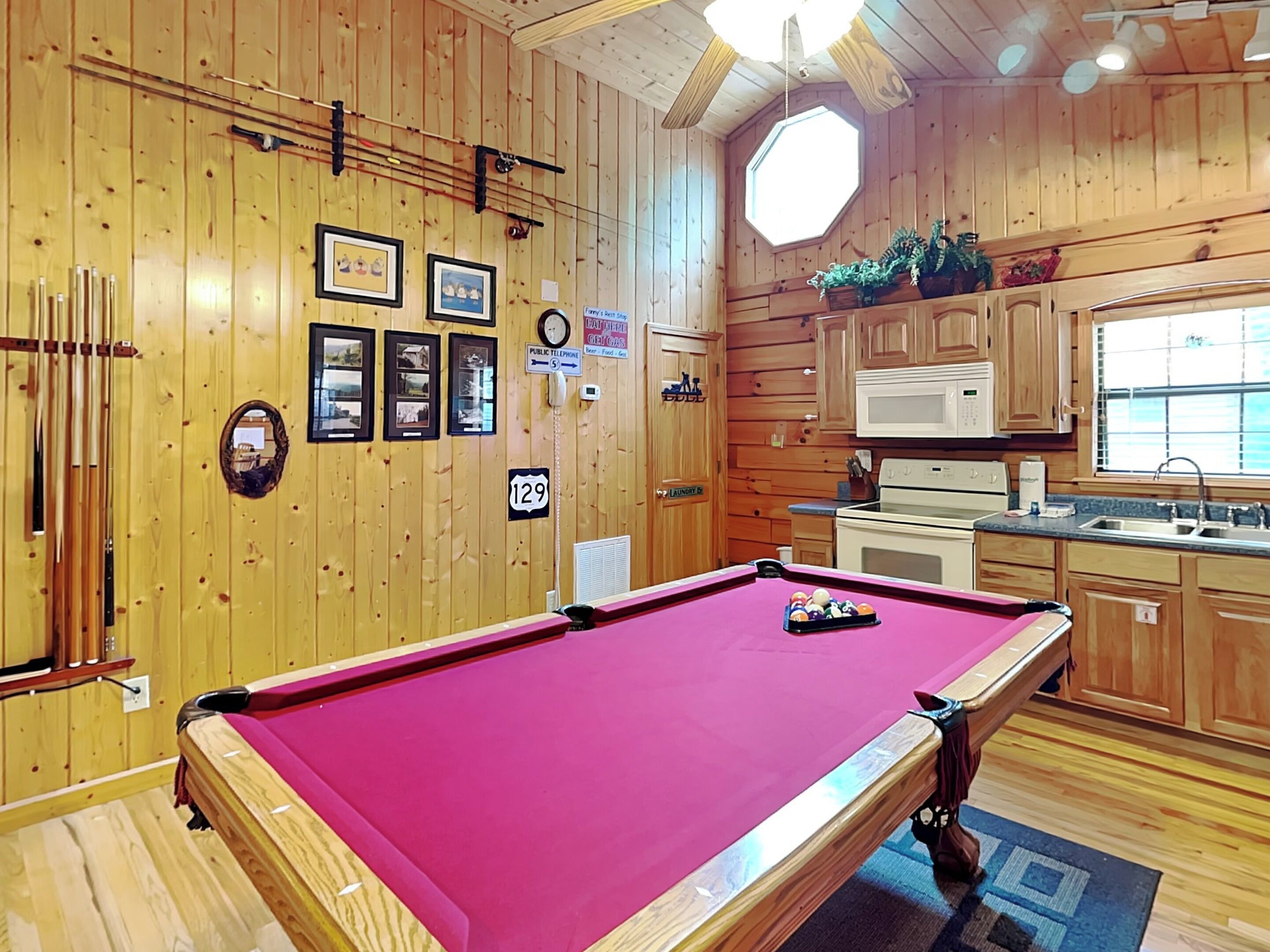 A pool table in a room with wooden walls, a clock, framed pictures, and a public telephone.