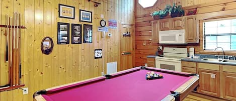A pool table in a room with wooden walls, a clock, framed pictures, and a public telephone.