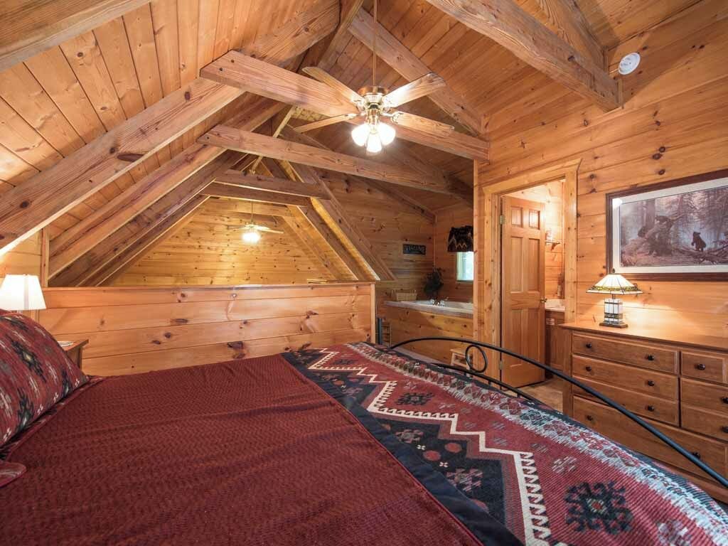 Un dormitorio acogedor en el ático, con techo inclinado, vigas de madera, cama con colcha estampada, escritorio con lámpara y un cuadro en la pared.