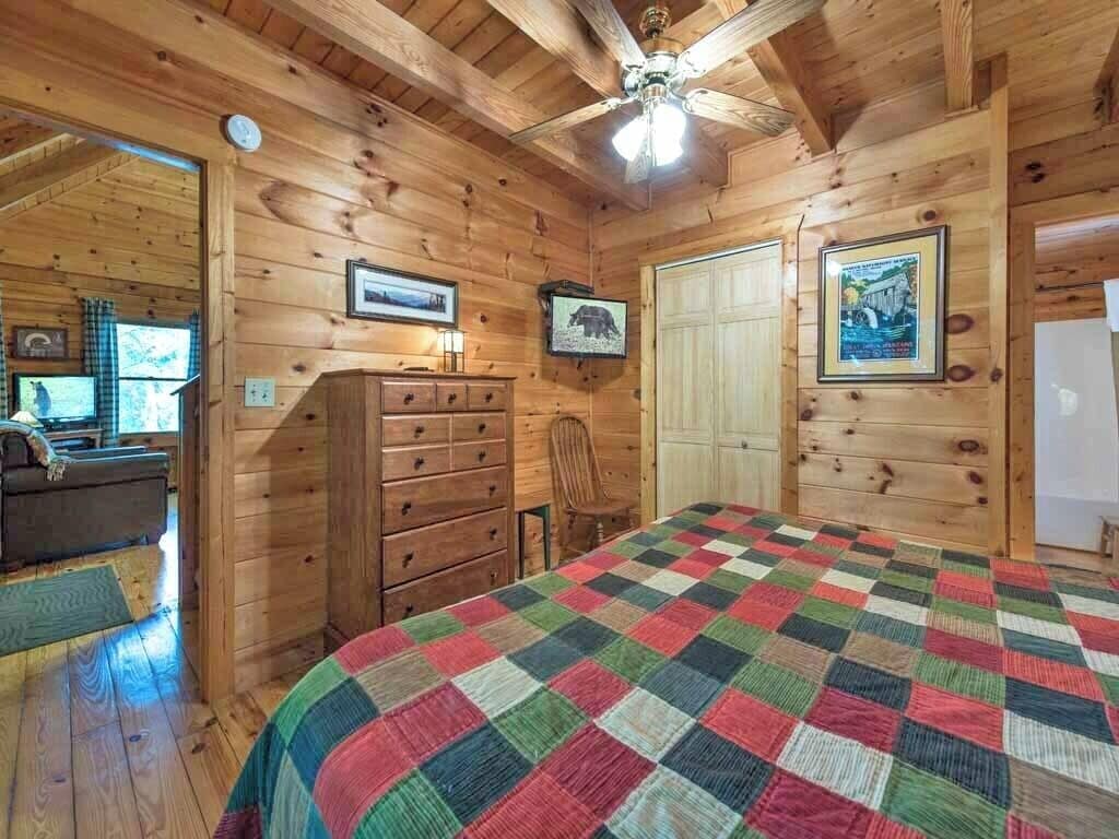 A wooden cabin bedroom with a quilted bedspread, wooden walls, a ceiling fan, and a dresser.