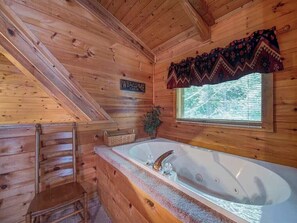 A cabin interior with a wooden interior, a large bathtub, a window with blinds, and a welcome sign. - Bear's Den by Jackson Mountain Rentals (Gatlinburg)