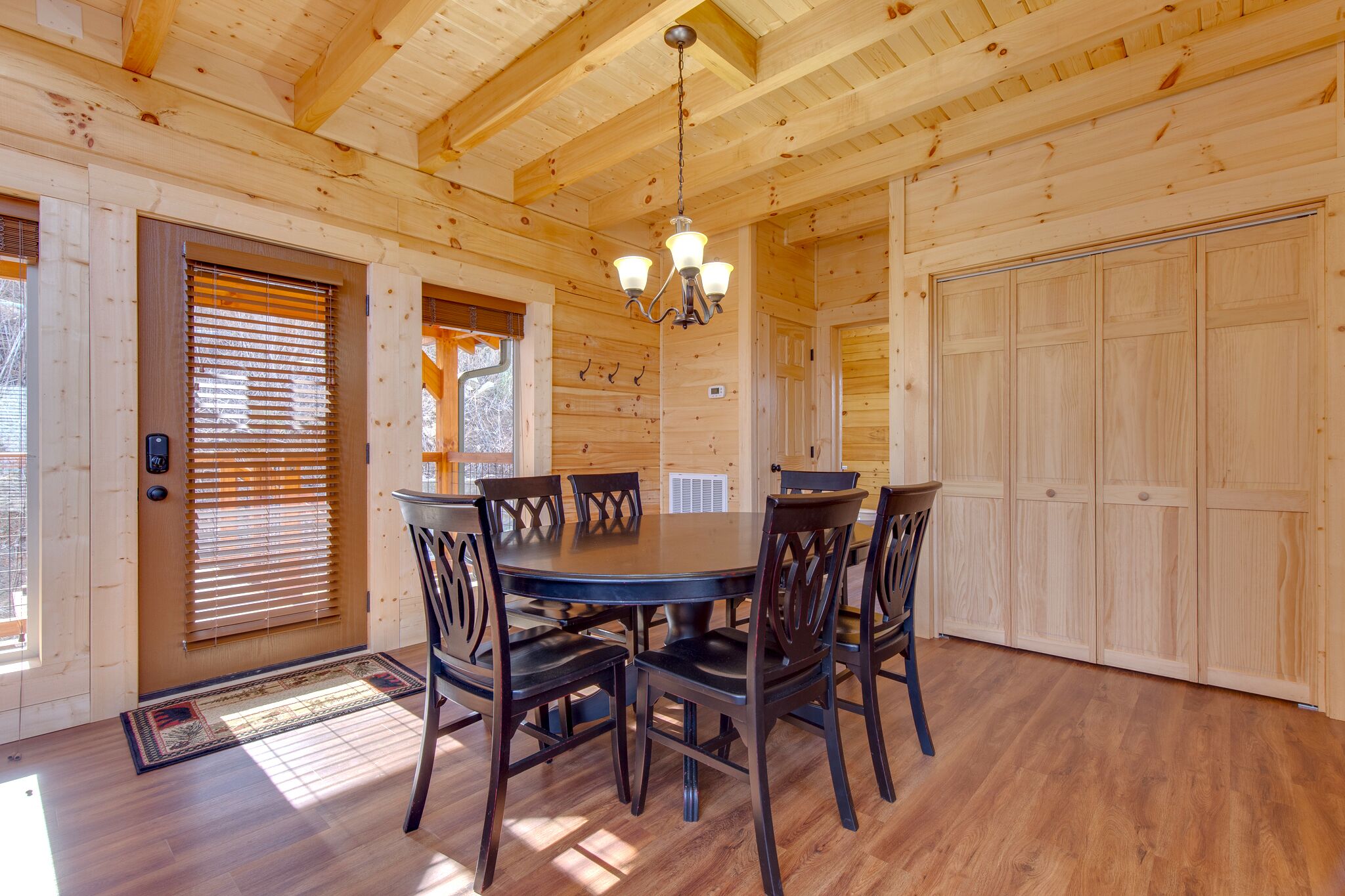 A wooden interior with a round dining table and chairs, a chandelier, and a large wooden door.