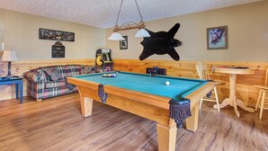 Una mesa de billar en una habitación con suelo de madera, un sofá escocés, una mesa de madera y una cabeza de oso montada.