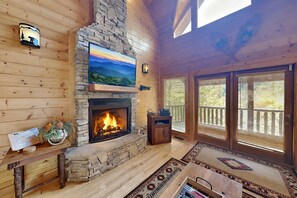 Interno di una baita in legno con camino in pietra, televisore a parete e balcone con ringhiera in legno.