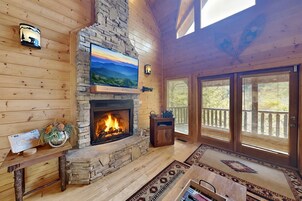 Interior de una cabaña de troncos con chimenea de piedra, televisor montado y balcón con barandilla de madera.