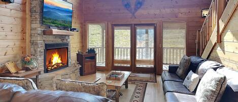 Un acogedor interior de cabaña con chimenea de piedra, sofás de cuero, un televisor grande y una vista panorámica a través de las ventanas.
