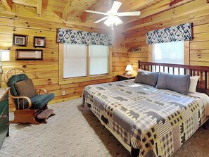 A wooden cabin bedroom with a bed, chair, nightstand, lamp, and two windows with shutters.