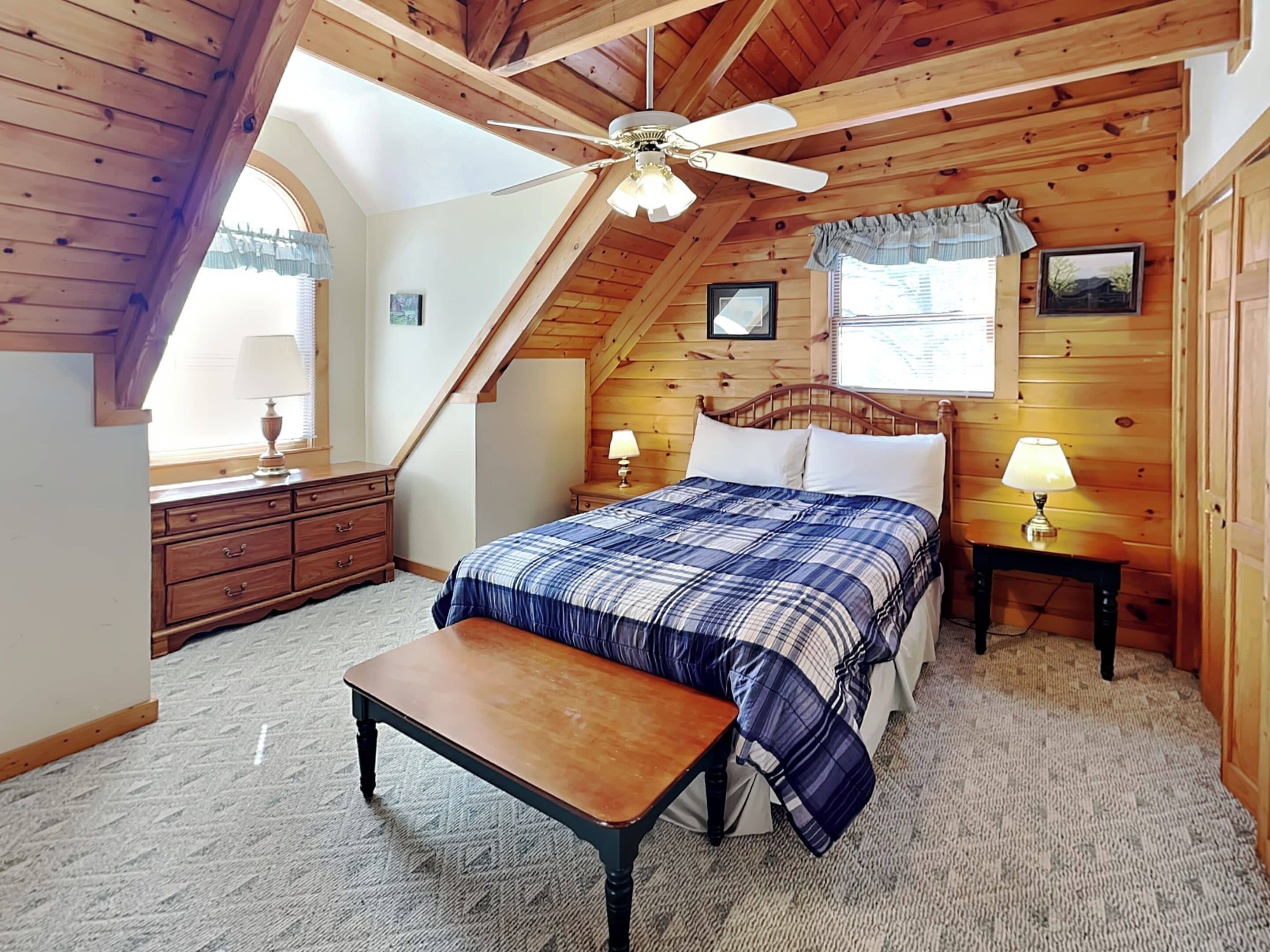 A bedroom with a wooden ceiling, a bed with a plaid bedspread, a nightstand with a lamp, a dresser, and a window with curtains.