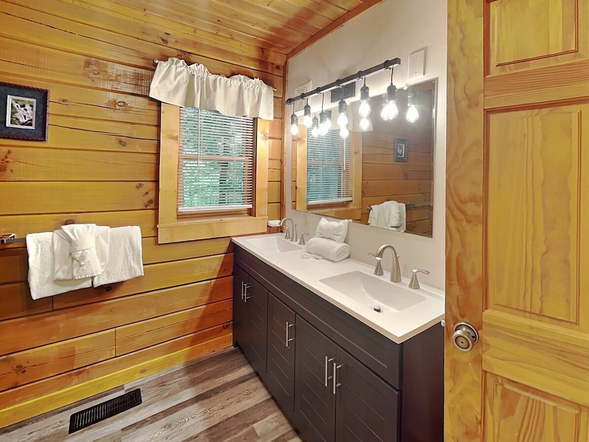 A wooden cabin bathroom with a large mirror, two sinks, and a window with blinds.