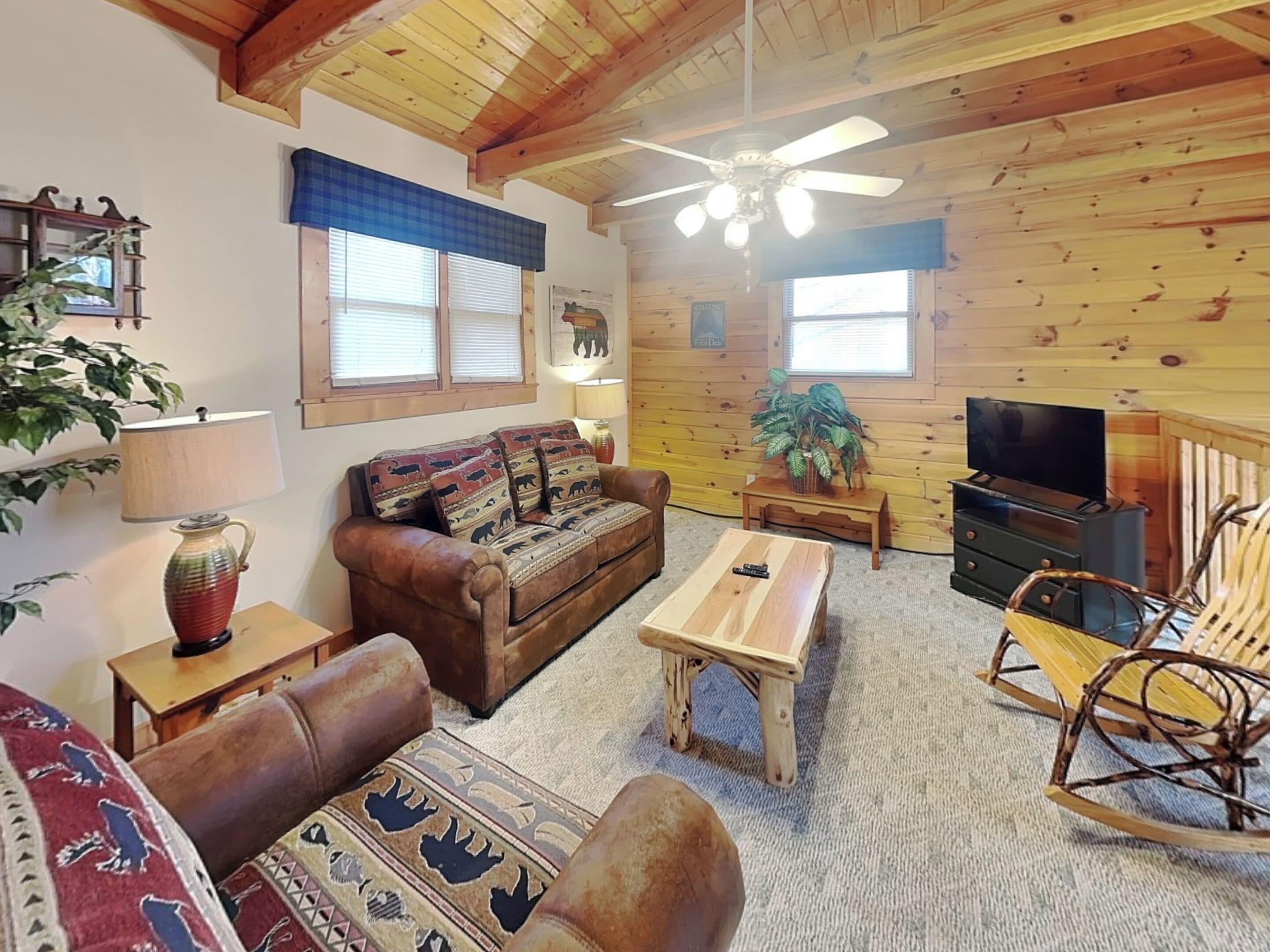 A cozy living room with a wooden ceiling, a brown leather sofa, a wooden coffee table, a rocking chair, and a television.