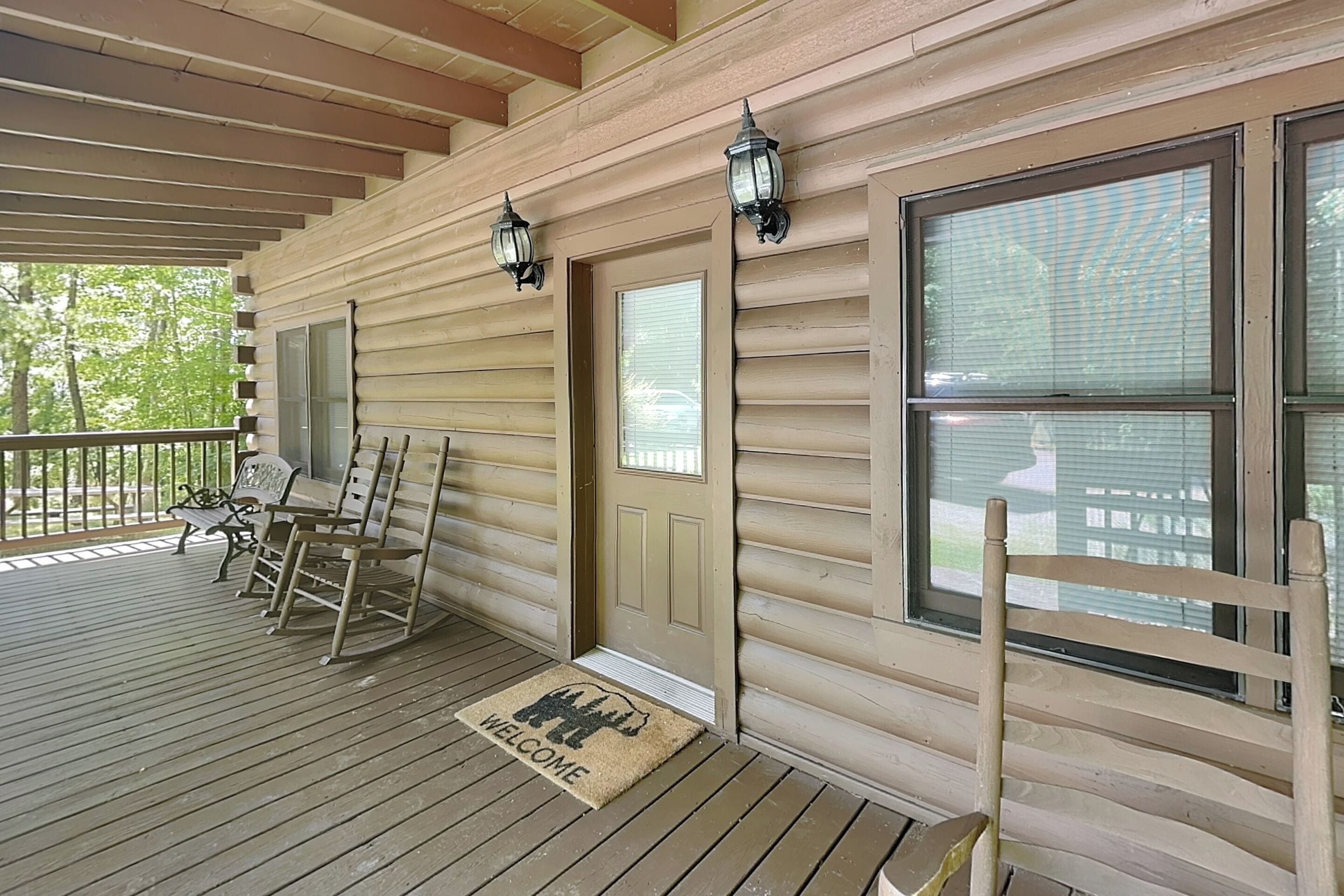 A wooden cabin with a porch, rocking chairs, a welcome mat, and two wall-mounted lanterns.