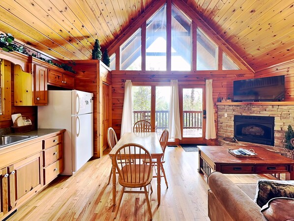 A cabin interior with wooden walls, a dining area, a kitchen with a refrigerator, and a fireplace. - Cub's Cove by Jackson Mountain Rentals (Gatlinburg)