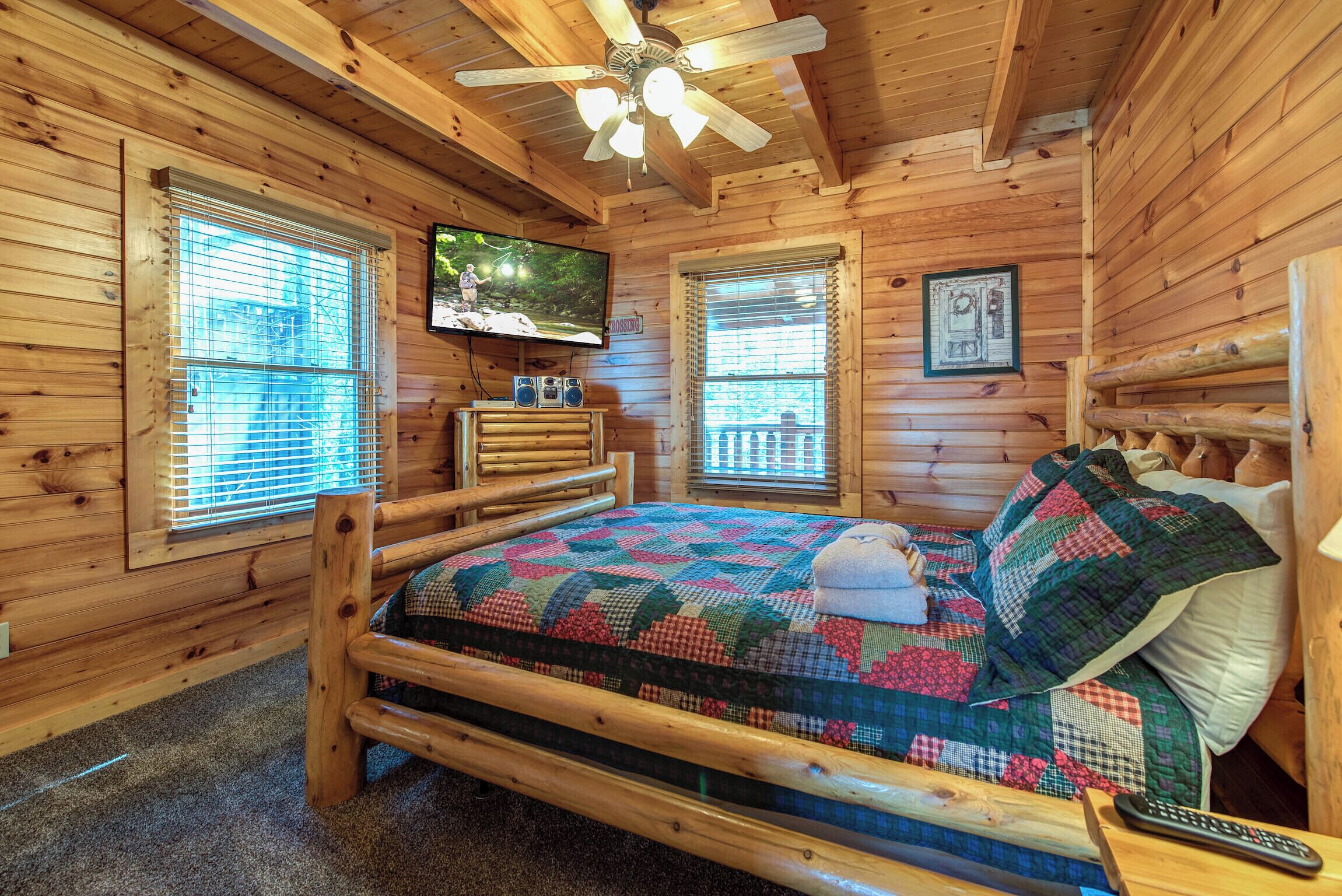 A wooden cabin room with a bed, a ceiling fan, a TV, and a window with blinds.