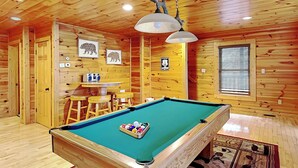 A pool table in a wooden room with a ceiling fan, a small bar area, and a window with blinds. - Cloud Dancer by Jackson Mountain Rentals (Gatlinburg)