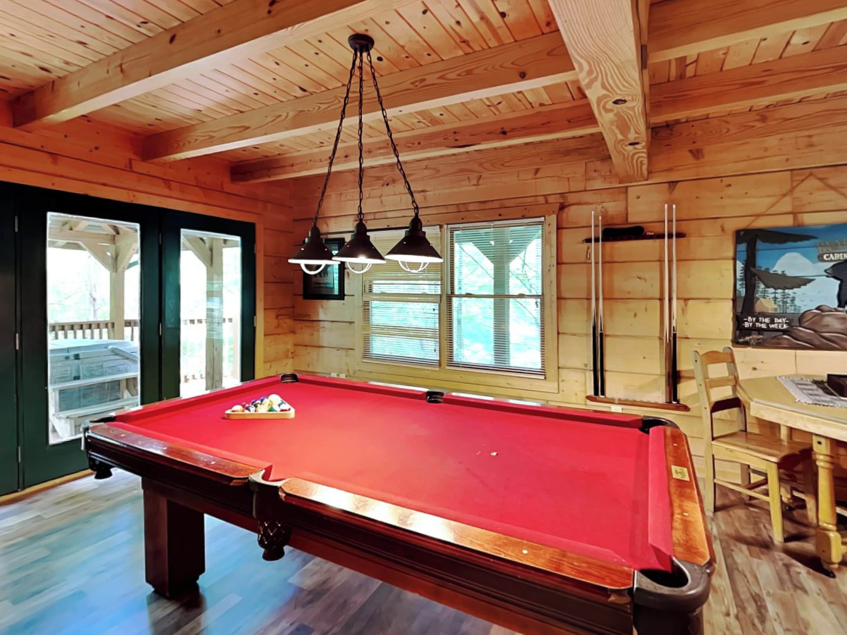 Une table de billard dans une pièce aux murs et au plafond en bois, une fenêtre avec des stores et une affiche accrochée au mur.