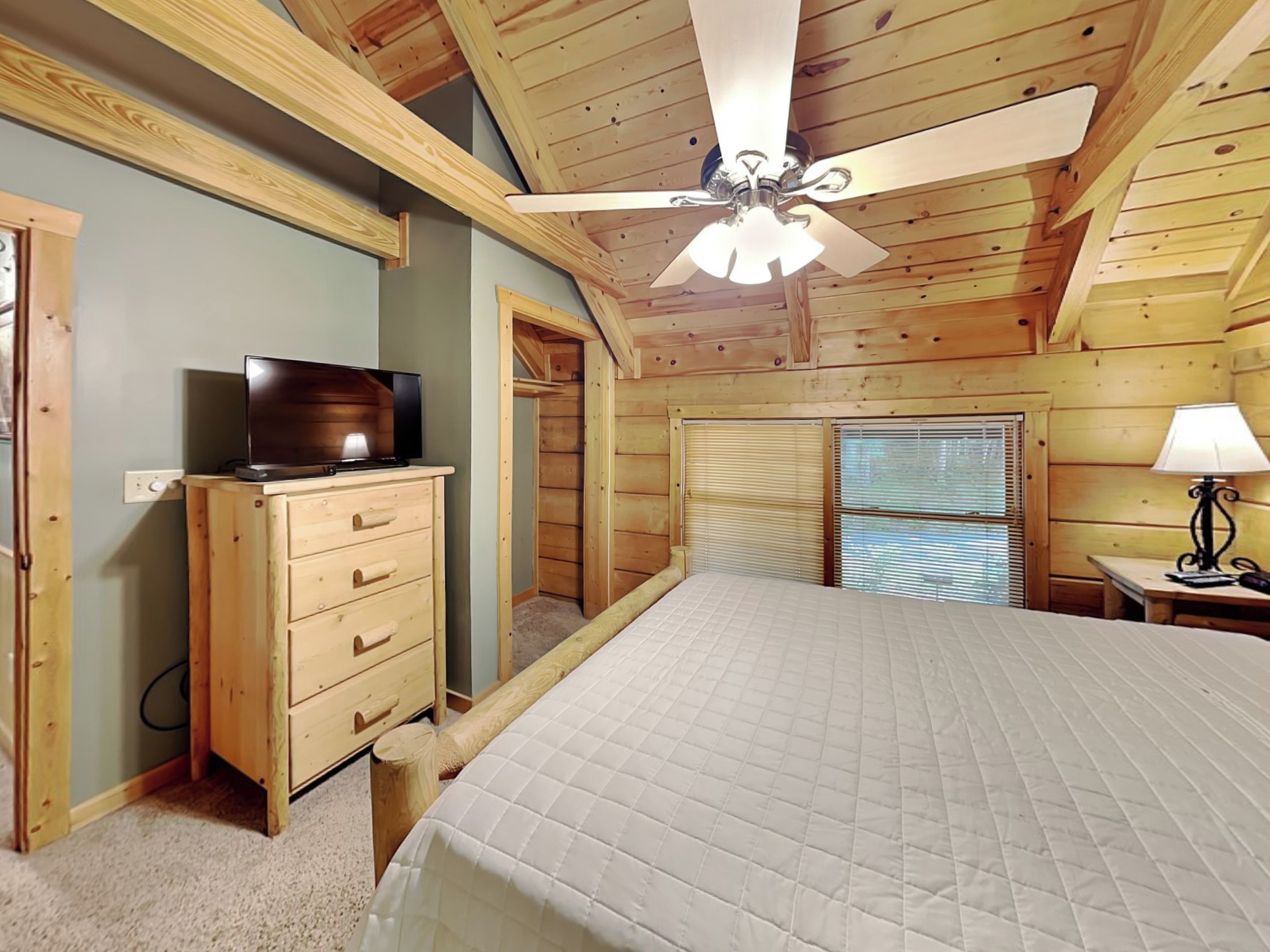 A bedroom with a bed, a ceiling fan, a TV on a dresser, a lamp, and a window with blinds.