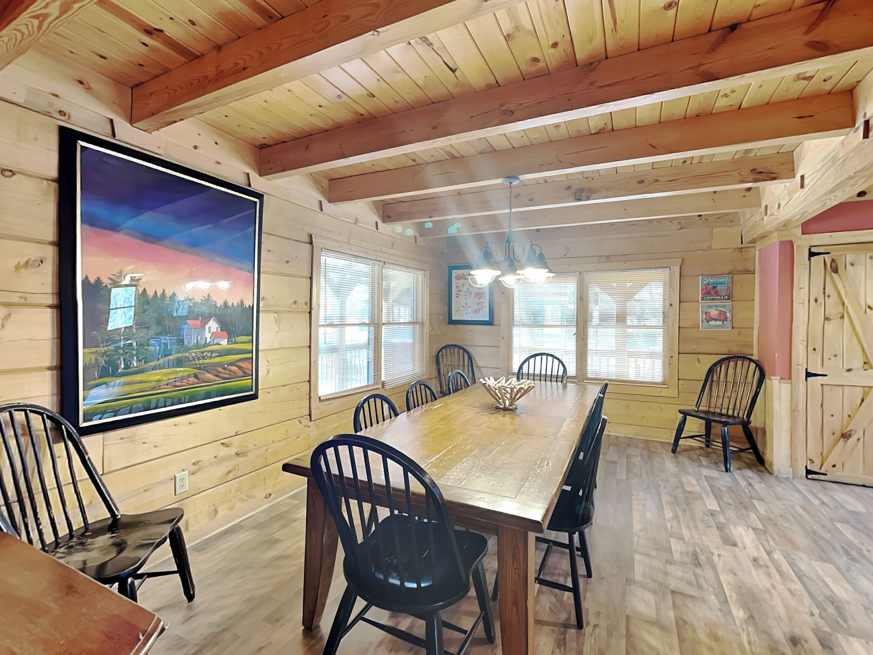 A dining area with a long wooden table, black chairs, and a painting of a landscape on the wall.