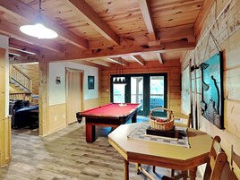 A wooden interior with a pool table, a dining table with a basket, and a staircase leading to another room.