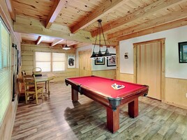 A pool table in a room with wooden floors and walls, a dining table with chairs, and a door.