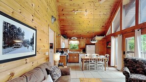A wooden cabin interior with a living room, dining area, kitchen, and large windows.