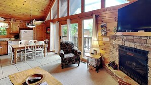 A cozy cabin interior with a dining area, a patterned armchair, a fireplace, and a television.