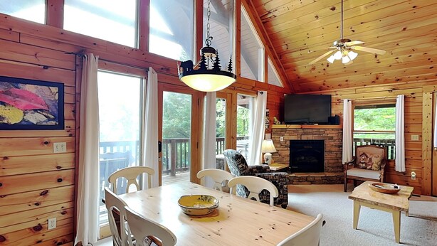 A wooden cabin interior with a dining table, chairs, a fireplace, a television, and large windows.