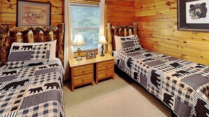 A cabin-style room with two beds, wooden walls, a window with curtains, and a bear-themed bedspread.